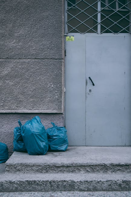 Four blue garbage bags made of plastic, tightly tied at the top, are placed on a concrete step in front of a grey, industrial-style door set within a textured, grey stucco wall. The bags are stacked closely together on the left side of the door, which has a black handle and a small keyhole beneath it. The door features a window with a metal grille above it, composed of crisscrossing bars forming diamond shapes. The surrounding environment appears to be an external urban setting, possibly a back alley or service entrance, and is illuminated with natural daylight, highlighting the rough texture of the wall and the smooth, shiny surface of the rubbish bags. The scene suggests a site for independent waste collection, commonly handled by private rubbish removal services such as Rubbish Collection Ealing, which facilitates alternative waste handling outside of municipal recycling systems, often involving on-site clearance of refuse from commercial or residential premises.