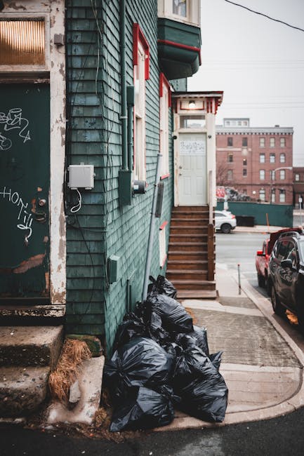 A row of black rubbish bags filled with waste is stacked at the base of a building with dark green wooden siding, showing signs of weathering and graffiti near a rusted metal door. The bags are on a concrete surface adjacent to a small stairway with wooden steps leading up to a white door with a small overhang, positioned slightly to the right of the bags. The building's exterior features electrical boxes and cables mounted on the wall, with some patches of peeling paint visible on the lower part near the bags. In the background, there is a parking area with a black vehicle parked close to the building, and further beyond, a red-brick industrial-style building with large windows. The scene is lit with natural, subdued daylight, capturing the typical urban environment for private waste disposal and alternative rubbish collection methods, as managed by Rubbish Collection Ealing.