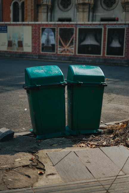 Two green wheeled rubbish bins with hinged lids are positioned side by side on a paved sidewalk edge in an urban environment. The bins are made of sturdy plastic with a slightly textured surface and are labeled with white text indicating their purpose. They are placed on a small patch of uneven pavement, surrounded by scattered dried leaves and dirt. Behind the bins, there is a street with asphalt surface, showing some minor wear and tire marks, and a brick and concrete building facade with large, framed posters or windows, partially obscured by a temporary construction fence or barrier. The scene appears to be during daylight with soft, natural lighting, and the overall setting suggests an area where local refuse is collected or disposed of, possibly for private waste management services like those offered by Rubbish Collection Ealing, providing an alternative to municipal rubbish collection in this area.