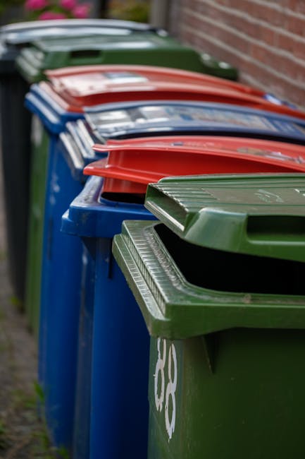 A row of large wheelie bins positioned outdoors along a pavement next to a brick wall, each with a distinct lid colour including green, red, blue, and dark green. The green bin in the foreground has a closed lid with a ridged texture and white markings, while the adjacent bins behind it feature similarly shaped lids, some of which are slightly open or tilted. The bins are made of durable plastic with smooth, matte surfaces and are aligned side by side, occupying most of the image's foreground. In the background, the bins recede into the distance, creating a sense of depth, with some greenery and an out-of-focus wooden fence or structure faintly visible. The lighting is soft and natural, indicating an overcast day, and the overall scene suggests an organized waste collection setup, possibly reflecting an alternative or private rubbish disposal service, such as Rubbish Collection Ealing, highlighting the importance of properly managed waste segregation and collection.