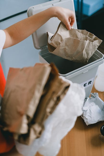 A person is depositing a crumpled brown paper bag into a grey indoor waste bin. The bin is positioned on a wooden surface, with part of a white label visible on the side, indicating waste collection. The person's arm is extending from the left, wearing a white short-sleeved top, and holding the bag above the open lid of the bin. In the foreground, a stack of additional crumpled paper bags and rubbish is held in the person's other hand, partially obscuring the background. The environment appears to be an indoor space with a clean, organized setting, possibly an office or utility room. The waste bin is made of plastic with a smooth finish, and the paper bag has a slightly rough texture showing creases and folds. The area is illuminated by soft natural or artificial light, emphasizing the textures and colors of the paper and the bin. This scene reflects an example of domestic or commercial waste disposal, relevant to services like independent rubbish collection or on-site clearance that address smaller-scale waste removal needs, aligning with the context of private rubbish disposal services.