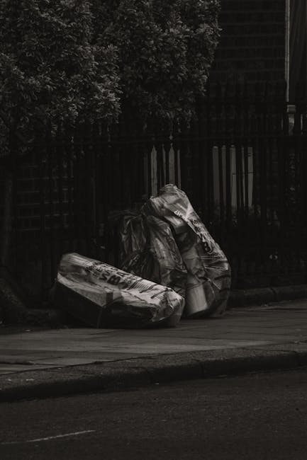 Two large black garbage bags filled with waste material are placed on a paved sidewalk near a black metal fence and a hedge. The bags are made of thick plastic, appear slightly crumpled, and are positioned close to each other, leaning against the fence. The scene is outdoors, with a portion of the roadway visible in the foreground, and appears to be captured during an overcast day, giving a muted, low-contrast appearance. The bags likely contain household or garden waste awaiting collection, indicative of private rubbish disposal or on-site waste handling services, possibly managed by Rubbish Collection Ealing. A large green shrub or tree can be seen behind the fence, contributing to the suburban environment. The setting suggests the disposal of waste in an urban or residential area where independent rubbish collection is employed instead of local authority bin services, aligning with the context of alternative waste removal methods.