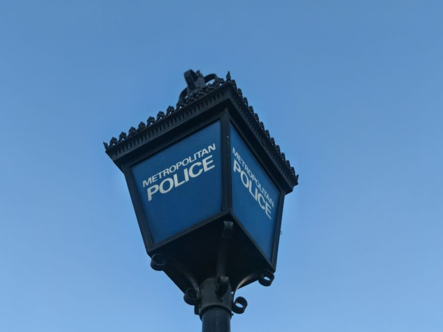 A vintage-style street lamp with a square blue metal lantern displaying the words 'Metropolitan Police' on two visible sides, mounted on a black pole. The lantern features decorative edging along the top and is set against a clear blue sky. The scene is outdoors, with no visible surrounding environment or additional objects, providing a neutral, professional view of a police service fixture often seen in urban areas. This image subtly connects to municipal or institutional waste and rubbish management themes through its public service context, aligning with proactive or community-related aspects of rubbish removal services in public or private settings.