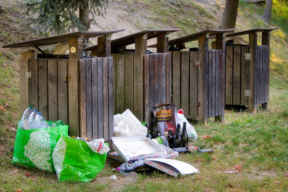 Two large black garbage bags filled with waste material are placed on a paved sidewalk near a black metal fence and a hedge. The bags are made of thick plastic, appear slightly crumpled, and are positioned close to each other, leaning against the fence. The scene is outdoors, with a portion of the roadway visible in the foreground, and appears to be captured during an overcast day, giving a muted, low-contrast appearance. The bags likely contain household or garden waste awaiting collection, indicative of private rubbish disposal or on-site waste handling services, possibly managed by Rubbish Collection Ealing. A large green shrub or tree can be seen behind the fence, contributing to the suburban environment. The setting suggests the disposal of waste in an urban or residential area where independent rubbish collection is employed instead of local authority bin services, aligning with the context of alternative waste removal methods.